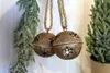 Two rustic, snowflake-cutout bells hang from twine, with a frosted evergreen garland on the left and a small potted evergreen tree on the right. Náhľad obrázku produktu