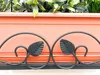 A close-up view of a black metal decorative scrollwork attached to an orange planter box filled with green artificial plants. Produktbild-Vorschau