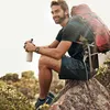 A man is sitting on a rock outdoors, holding a water bottle and wearing a backpack. Náhled obrázku produktu