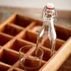 A small glass bottle with a swing top closure and a small glass sit next to each other in a wooden grid organizer. Produktbild-Vorschau