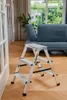 A step ladder is placed on a wooden floor in a living room setting, with a sofa and curtains in the background. Produktbild-Vorschau