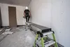 A construction worker stands on a telescopic plank in an unfinished room. Produktbild-Vorschau