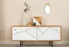 A modern sideboard with white doors featuring geometric gold lines, a wooden top, and black legs, decorated with a vase of greenery, a small potted plant, and a framed artwork. Produktbild-Vorschau