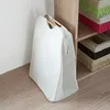 A white laundry bag with wooden handles stands on a wooden floor next to a shelf unit filled with storage baskets. Produktbild-Vorschau