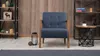 A single blue armchair is placed in a room with a wooden wall, a bookshelf, and a side table. Produktbild-Vorschau