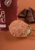 A close-up shot of cocoa powder in a measuring spoon, with a can of the product and pieces of dark chocolate in the background. Voir les images du produit