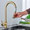 A hand is shown using a gold kitchen faucet with a pull-down sprayer over a sink. Cucumbers and lettuce are visible on a cutting board in the background. Náhled obrázku produktu