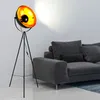 A black tripod floor lamp with a gold interior is positioned next to a dark gray sofa in a room. Produktbild-Vorschau