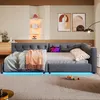 A modern bedroom or living room setting featuring a gray upholstered daybed with LED lighting underneath. The daybed is made up with a light gray duvet and a pink throw blanket. A tablet is placed on the daybed, and decorative items are visible on shelves  Produktbild-Vorschau