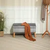 A grey upholstered storage bench with a brown throw blanket draped over it, placed in a room with paneled walls and a coat rack in the background. Náhled obrázku produktu