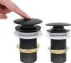 A hand is pressing down on a black drain stopper, with another identical drain stopper to the right. Produktbild-Vorschau
