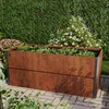 A raised garden bed made of corten steel with a rust-like appearance, filled with various plants and herbs, situated on a paved patio in a garden setting with a lush green hedge and flowering bushes in the background. Produktbild-Vorschau