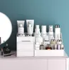 A white cosmetic organizer filled with various skincare and makeup products is placed on a light pink surface in front of a teal blue wall. A round mirror is partially visible on the left. Several bottles, jars, tubes, and a perfume bottle are arranged nea Náhľad obrázku produktu