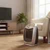 A ceramic heater is placed on a wooden floor in a living room next to a chair and a TV stand. Podgląd zdjęcia produktu
