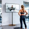 A woman is doing yoga in front of a TV screen displaying a yoga tutorial. The TV is mounted on a floor stand. Anteprima dell’immagine prodotto