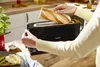 A person is putting slices of bread into a black toaster on a wooden kitchen counter. Produktbild-Vorschau