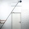 A person is using a long-handled brush to clean an air conditioner unit mounted on a white wall. A white door is visible in the background, along with a tiled wall. Podgląd zdjęcia produktu