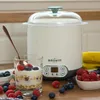 A yogurt maker is shown on a wooden surface with a glass of yogurt and berries in the foreground. Náhled obrázku produktu