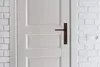 A close-up view of a white door with a brown door handle, set against a white brick wall. Anteprima dell’immagine prodotto
