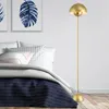 A golden floor lamp stands next to a bed in a bedroom. Voir les images du produit
