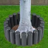 A tree trunk surrounded by a ring of dark gray garden edging, set against a backdrop of green grass and a textured wall. Voir les images du produit