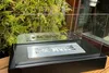 A black freestanding bio-fireplace with a glass enclosure sits outdoors on a wooden surface, with green foliage in the background. Produktbild-Vorschau