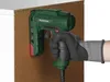 A hand wearing a glove is holding a green electric tacker against a brown surface, possibly demonstrating its use. Produktbild-Vorschau