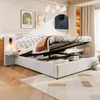 A bedroom scene featuring a white upholstered storage bed with its hydraulic lift mechanism open, revealing storage space underneath. A bedside table with a lamp is attached to the headboard, and decorative artwork hangs on the wall. Voir les images du produit