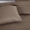 A close-up view of a brown quilted blanket with a geometric triangle pattern, showing a pillow in the foreground. Produktbild-Vorschau