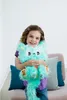 A young girl is holding a fluffy teal toy creature while sitting on a couch. Produktbild-Vorschau