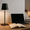 A black desk lamp, an open book, and a laptop are placed on a wooden desk. Produktbild-Vorschau