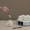 A hand is holding a spoon over a glass jar of yogurt, with a yogurt maker and more jars in the background. Produktbild-Vorschau