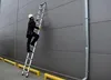 A person is climbing an aluminum telescopic ladder against a gray building wall. Náhľad obrázku produktu