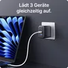 A laptop is partially visible on the left, connected to a black GaN charger plugged into a wall outlet on the right. The charger has two USB-C ports and one USB-A port, with a white cable connecting it to the laptop. Produktbild-Vorschau