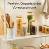 A shelf displaying various glass storage jars filled with dry goods like pasta, grains, and spices, along with wooden kitchen utensils. A tea towel hangs in the background. Produktbild-Vorschau