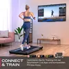 A woman is running on a treadmill in a living room while looking at a large TV screen displaying a running simulation. The scene includes furniture and plants, suggesting a home fitness setup. Produktbild-Vorschau