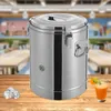 A stainless steel insulated container sits on a wooden table, with ice cubes and a glass of juice in the foreground. The background shows a blurred view of a dining hall or cafeteria. Produktbild-Vorschau