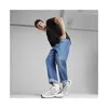 A man wearing a black t-shirt and blue jeans is posing in a studio setting, leaning forward with his hands in his pockets, showcasing his white and black Puma sneakers. Voir les images du produit
