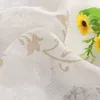 A close-up of white sheer fabric with a gold floral pattern, with two yellow sunflowers and green leaves in the background. Produktbild-Vorschau