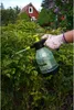 A hand wearing a glove is holding a spray bottle and spraying a plant in a garden setting. A red house is visible in the background. Anteprima dell’immagine prodotto