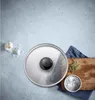 A glass pot lid is shown from a top-down view on a light gray textured surface, with ingredients like garlic and salt scattered around. Podgląd zdjęcia produktu