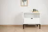 A white nightstand with a drawer and an open shelf sits on a light wooden floor against a white wall. A framed picture hangs on the wall above the nightstand, and a wooden tray with two green glasses sits on top of the nightstand. Produktbild-Vorschau