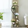 A multi-tiered plant stand with various floral arrangements is placed near a window with sheer curtains and a sofa. Anteprima dell’immagine prodotto