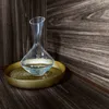 A clear glass decanter filled with a light-colored liquid sits on a textured gold tray, placed against a dark wood-paneled background. Náhľad obrázku produktu