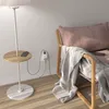 A smart plug is shown plugged into a wall outlet, with USB cables connected to a phone on a side table next to a floor lamp. A blanket is draped over a chair in the background. Náhľad obrázku produktu