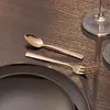 A close-up shot of a bronze-colored spoon and fork set placed on a dark wooden table next to a black plate. Náhľad obrázku produktu