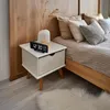 A bedside table with a digital clock, vase, and a bed in the background. Produktbild-Vorschau