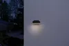 A dark gray outdoor LED light fixture is mounted on a textured light gray wall. The light is on, casting a warm glow downwards. To the left, a dark, out-of-focus background shows trees and a building. Produktbild-Vorschau