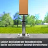 A close-up of a metal post support with a wooden post attached, set against a blurred background of a grassy field and trees under a blue sky. Produktbild-Vorschau