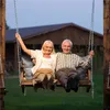 An elderly couple sits on a swing in front of a wooden house. Náhled obrázku produktu